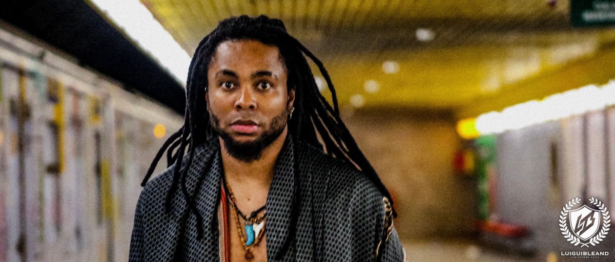 Colour photograph of Luigui Bleand walking toward the camera through an underground corridor, in a dark patterned jacket and necklaces, the corridor behind him out of focus