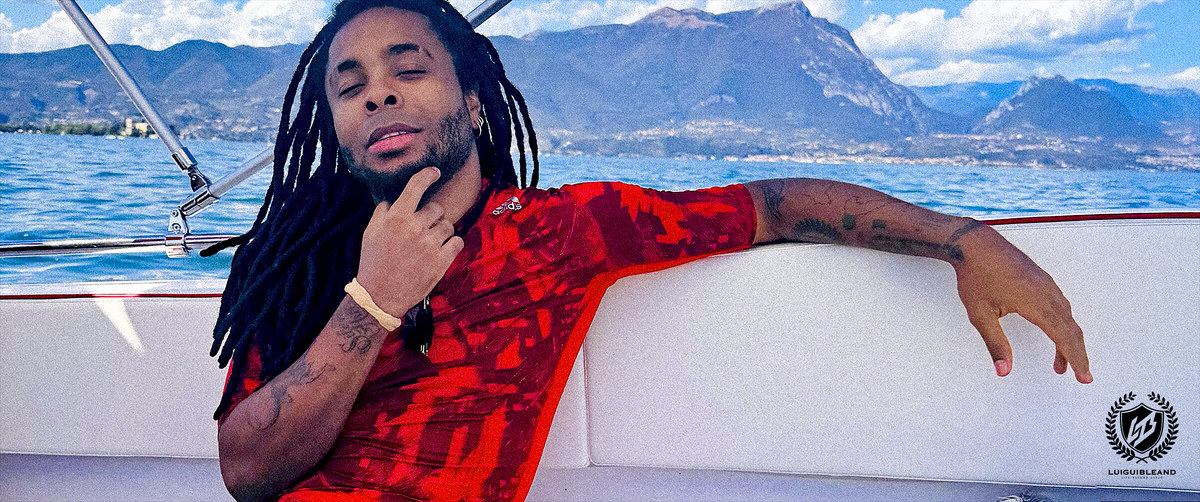 Luigui Bleand sitting on a boat with one arm along the backrest, water and mountains behind him; his crest in the corner