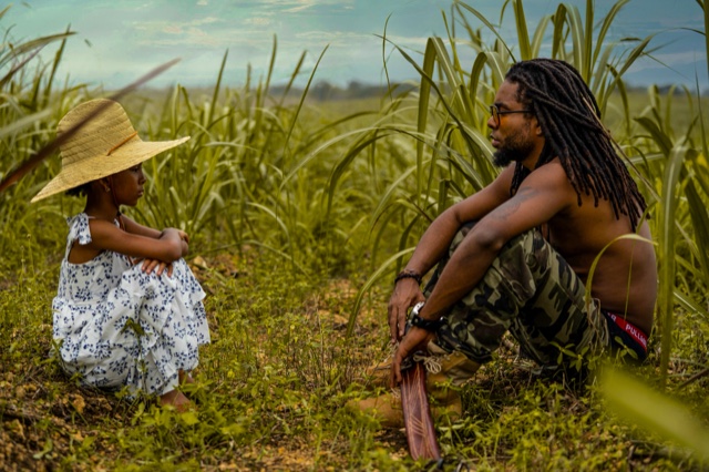 Colour photograph of Luigui Bleand sitting among tall sugarcane, facing Allyson, a young girl in a straw hat and a floral dress, under a cloudy sky